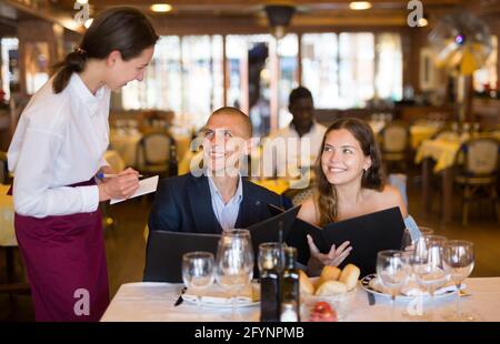 Cameriera cordiale e sorridente che consulta la coppia di ospiti che scelgono bevande e pasti al ristorante Foto Stock