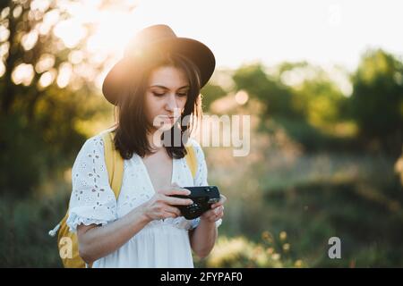 Look alla moda, bella cool giovane donna modello con fotocamera retrò pellicola e indossare un cappello elegante, vestito bianco, zaino giallo. VIN dell'azienda turistica femminile Foto Stock