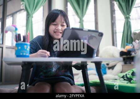 Piccola studentessa asiatica studiando durante la sua lezione in linea a casa con sorriso e felice, distanza sociale durante quarantena, concetto di educazione in linea Foto Stock