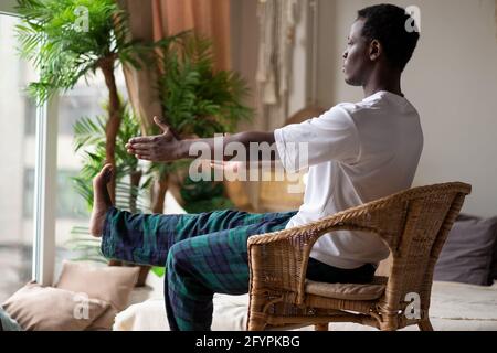 Uomo africano che usa la sedia nel suo soggiorno a casa facendo yoga asana per i principianti. Foto Stock