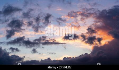 Il suggestivo e colorato cielo del tramonto con il sole che illumina parte del nuvole che li dipingono un bagliore arancione contro un cielo blu con macchie scure in primo piano Foto Stock