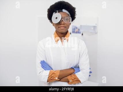 Ritratto di giovane donna afro-americana piacevole, fiducioso medico otolaringologo ENT, indossando camice da laboratorio bianco, riflettore frontale della testa e occhiali protettivi, in posa a macchina fotografica con le braccia incrociate Foto Stock