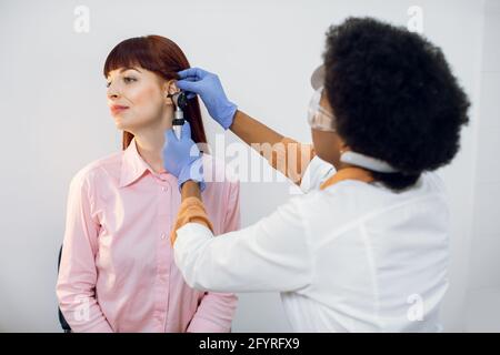 Concetto di diagnosi di perdita dell'udito. Primo piano di femmina giovane dottore otorhinolaringologo che guarda pazienti orecchio con otoscopio. Giovane donna afroamericana che ha controllato in su del medico di ENT. Foto Stock