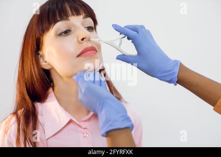 Primo piano del volto di una giovane ragazza dai capelli rossi durante l'esame medico nell'ufficio ENT. Mani di otolaringologo in guanti blu, che esaminano il naso di un paziente giovane con uno strumento monouso Foto Stock