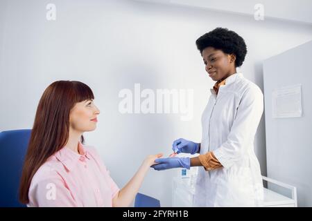 Tecnico di laboratorio afro-americano, trafilando sangue per le dita per il test. Giovane paziente caucasico signora fidarsi del suo medico e avere un esame del sangue Foto Stock