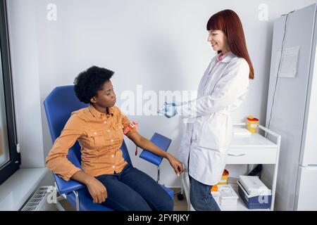 Analisi del sangue per donne in gravidanza. Bella giovane donna afroamericana seduta al laboratorio moderno, mentre piacevole giovane infermiera caucasica mostra la sua provetta con il campione di sangue Foto Stock