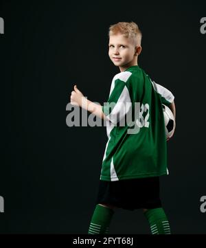 Bambino biondo sorridente in abiti sportivi verdi e bianchi in piedi tenendo la palla di calcio e mostrando pollici in su segno Foto Stock