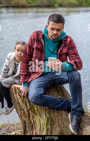 Papà sta controllando il suo telefono, non prestando attenzione a sua figlia, per una passeggiata nel bosco. Foto Stock