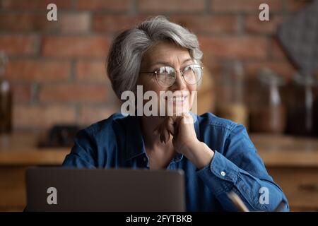 Donna anziana siede in cucina vicino il sorriso del laptop sguardo via Foto Stock