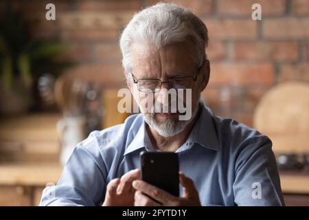 Uomo serio maturo in occhiali sedersi in cucina che tiene smartphone Foto Stock