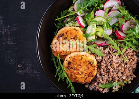 Cena sana. Ciotola per il pranzo con porridge di grano saraceno, cotolette di pollo fritte e insalata di arugula, cetriolo e rafano di verdure fresche. Vista dall'alto, appartamento la Foto Stock
