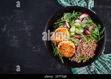 Cena sana. Ciotola per il pranzo con porridge di grano saraceno, cotolette di pollo fritte e insalata di arugula, cetriolo e rafano di verdure fresche. Vista dall'alto, appartamento la Foto Stock