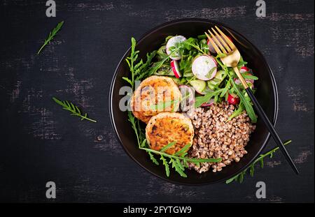 Cena sana. Ciotola per il pranzo con porridge di grano saraceno, cotolette di pollo fritte e insalata di arugula, cetriolo e rafano di verdure fresche. Vista dall'alto, appartamento la Foto Stock