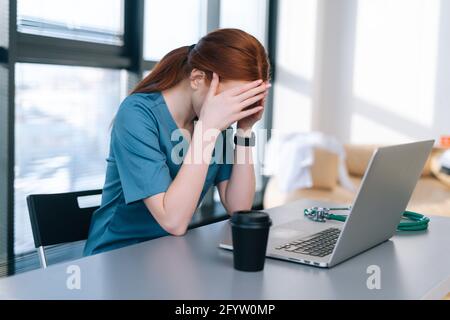 Vista laterale di stanco esausto giovane medico indossando medico uniforme che ha mal di testa e sfregamento testa con le mani Foto Stock