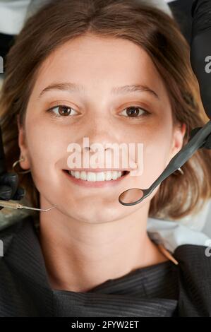 Vista dall'alto su una giovane ragazza sorridente che visita il dentista, sdraiata su una sedia dentista, indossando un bib nero. Strumenti dentista accanto al viso. Primo piano dell'istantanea. Concetto di igiene della bocca Foto Stock