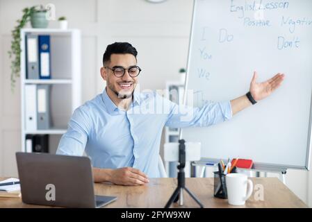 E-learning. Insegnante arabo maschile che dà online lezione di inglese su smartphone, puntando alla lavagna con le regole grammaticali Foto Stock