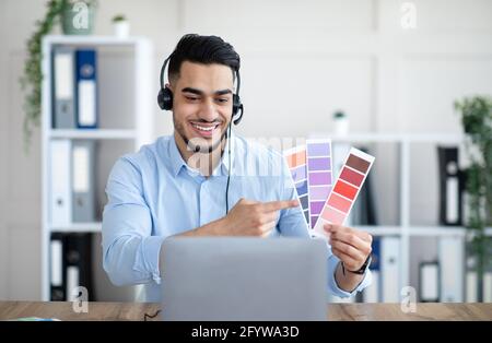 Uomo arabo che tiene campioni di colore davanti al laptop in ufficio, presentando un progetto di grafica durante la riunione online Foto Stock