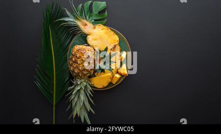 Ananas a fette su un piatto con foglie di palma tropicali. Bromelina intera ananas estate frutta metà ananas su sfondo nero scuro. Frutta estiva Foto Stock