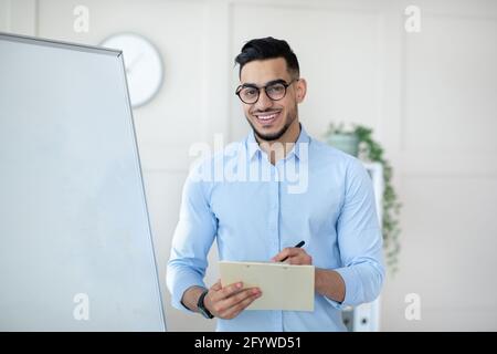 Moderna scuola online. Allegro insegnante arabo in piedi vicino a lavagna vuota con appunti, lezione di insegnamento sul web Foto Stock