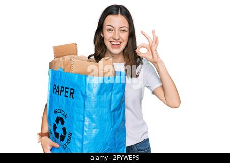 Giovane bella donna che tiene riciclaggio wastcanestro con carta e cartone facendo ok segno con le dita, sorridente amichevole gesturing simbolo eccellente Foto Stock