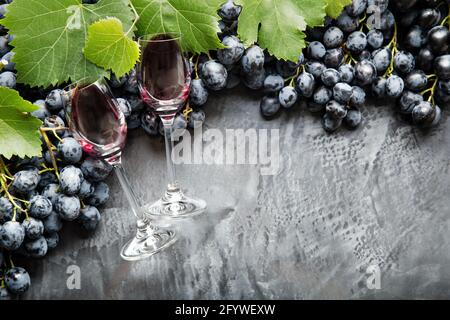 Due bicchieri di vino ripieni di vino sdraiati in cornice di uva. Uve nere succose vino rosso su fondo di cemento scuro d'annata. Spazio di copia su nero Foto Stock