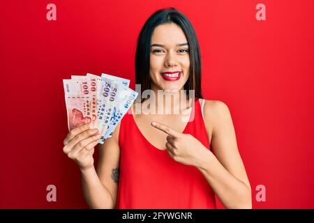 Giovane ragazza ispanica che tiene nuove banconote in dollari di taiwan sorridente felice puntare con la mano e il dito Foto Stock