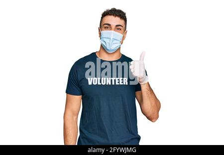 Giovane ispanico che indossa maschera medica e t shirt volontaria sorridente felice e positivo, pollice su facendo eccellente e segno di approvazione Foto Stock