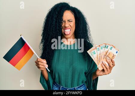 Donna afro-americana di mezza età che tiene la bandiera della germania e le banconote di euro che attaccano la lingua fuori felice con l'espressione divertente. Foto Stock