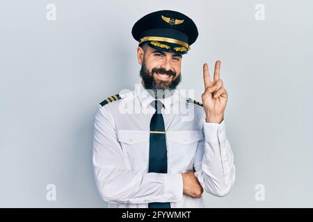 Giovane uomo ispanico che indossa l'uniforme pilota di aeroplano sorridente con felice faccia che si inala alla macchina fotografica facendo segno di vittoria. Numero due. Foto Stock