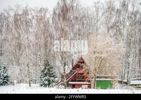 Bella casa di legno rosso nella foresta di fata di neve. Scenario invernale con casetta rossa circondata da alberi ricoperti di neve e gelo. Foto Stock