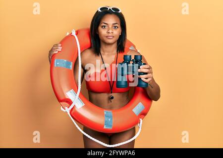 Giovane afroamericana ragazza che tiene salvagente galleggiante usando binocoli rilassato con espressione seria sul viso. Semplice e naturale guardare il venuto Foto Stock