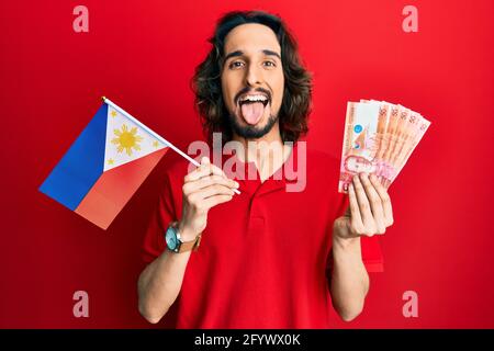 Giovane uomo ispanico che tiene bandiera filippine e pesos banconote che attaccano la lingua fuori felice con l'espressione divertente. Foto Stock