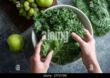 Le mani femminili lavano il cavolo di kale in ciotola di acqua. Cucina sana casa biologica coltivata vegan cibo, vista dall'alto Foto Stock