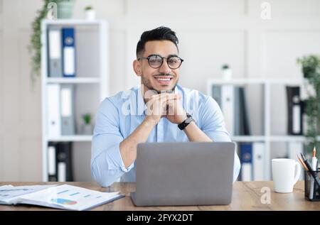 Ritratto di giovane arabo professionista seduto alla sua scrivania con un pc portatile in un ufficio moderno Foto Stock