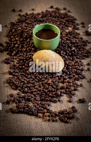 Concetto di colazione. Una tazza di caffè verde e un biscotto fatto in casa circondato da chicchi di caffè. Un tessuto di iuta sullo sfondo. Foto Stock