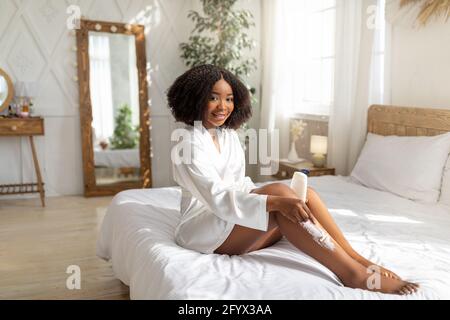 Lunghezza completa di signora nera che rade le gambe con rasoio, seduto sul letto in accappatoio, facendo la procedura di bellezza domestica Foto Stock