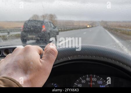 mano del conducente sul volante all'interno della vettura e gocce di pioggia sul parabrezza Foto Stock