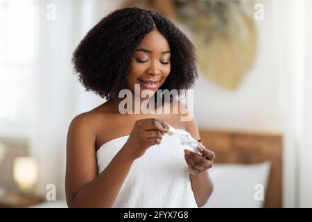 Attraente donna afroamericana con pelle sana e brillante applicando idratante siero facciale a casa Foto Stock