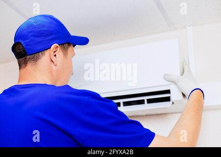 Un tecnico pulito in un cappello blu da baseball e tute fissa il nuovo condizionatore d'aria alla parete nella stanza. Ritratto di un lavoratore dalla parte posteriore. Derisione Foto Stock