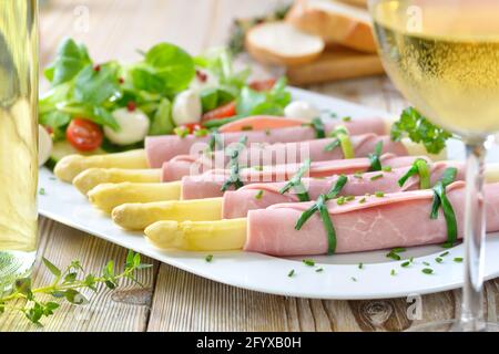 Deliziosa insalata di primavera con asparagi arrotolati in prosciutto cotto su un piatto bianco, servito con un vino bianco secco giovane e fette di baguette Foto Stock