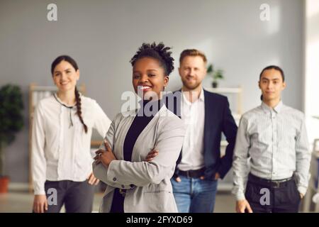 Donna d'affari afroamericana in piedi braccia piegate e sorridenti con la squadra di lavoratori dietro Foto Stock