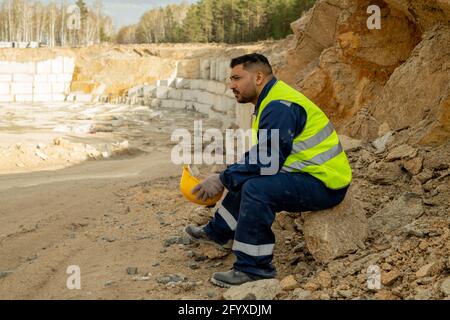 Medio vecchio costruttore in uniforme seduta su pietra mentre ha pausa Foto Stock