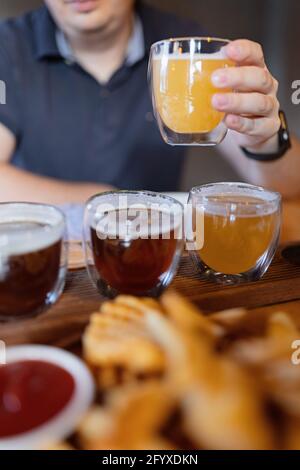 Uomo assaggiando varietà di birre artigianali stagionali in pub. Campionatori di birra in piccoli bicchieri posizionati singolarmente in fori realizzati in un unico vassoio di legno Foto Stock