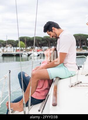 Vista laterale di una coppia multirazziale sorridente che parla mentre si guarda uno sull'altro in yacht nel porto della città Foto Stock