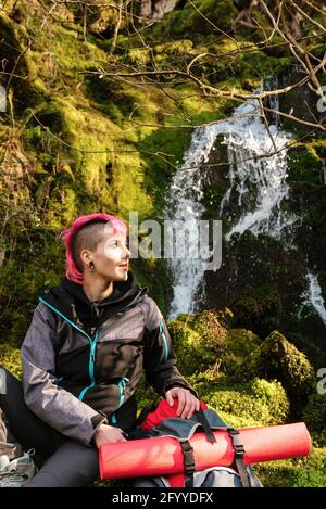 Donna escursionista con capelli tinti che guardano via mentre si siede sopra rocce mussose con zaino vicino alla cascata che cade dalla scogliera rocciosa Foto Stock