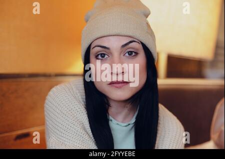 Giovane donna con occhi marroni in maglia e cappello guardando la fotocamera su sfondo sfocato Foto Stock