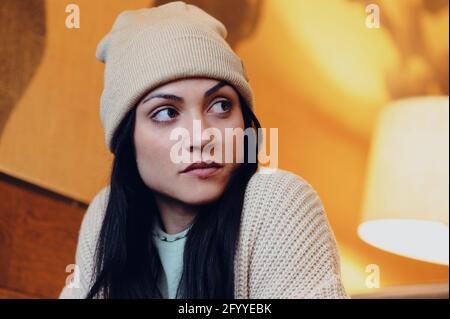 Giovane donna con occhi marroni in maglia e cappello guardando lontano su sfondo sfocato Foto Stock