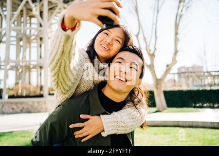 Amata giovane donna asiatica che abbraccia la schiena del ragazzo prendendo selfie su smartphone durante la data romantica nel parco Foto Stock