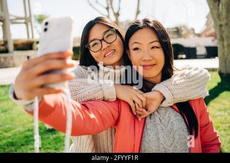 Amici giovani etnici positivi con lunghi capelli scuri Vestiti casual sorridente e mostrando il segno V mentre si prende selfie su smartphone il giorno di sole io Foto Stock