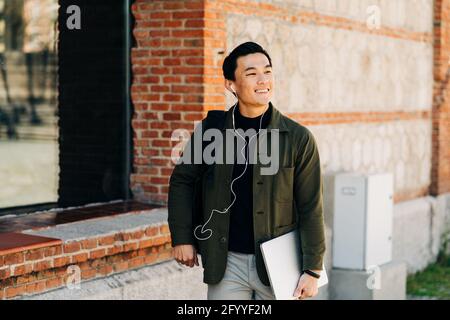 Positivo giovane etnico maschile millenario in elegante vestito sorridente mentre ascoltare la canzone preferita con gli auricolari che camminano lungo la strada della città con portatile in mano Foto Stock